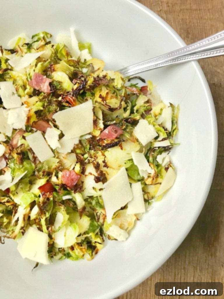 A large white serving bowl with a silver spoon on a wooden surface. In the bowl is crispy shaved brussels sprouts topped with diced turkey bacon, shaved parmesan cheese, and a balsamic vinegar reduction.
