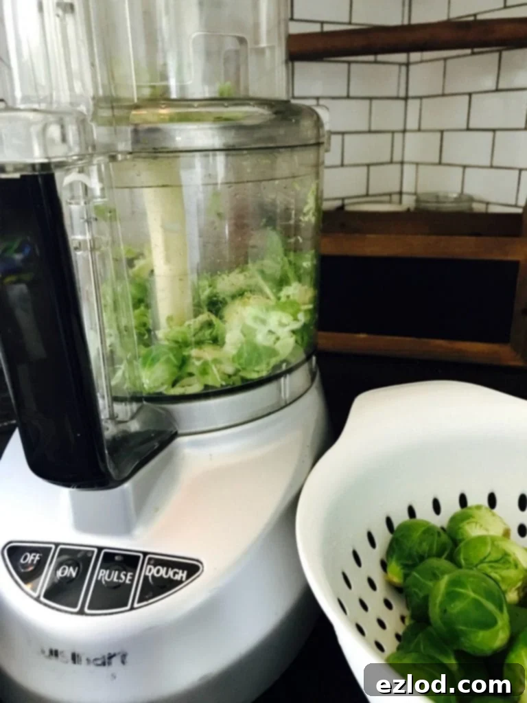 White Cuisinart food processor filled with already shaved brussels sprouts. On the side is a white colander with whole brussels sprouts. In the background there is a white kitchen backsplash and wooden shelf.
