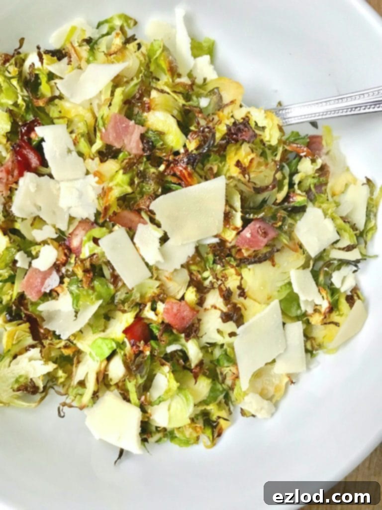 Close up of a large white serving bowl with a silver spoon on a wooden surface. In the bowl is crispy shaved brussels sprouts topped with diced turkey bacon, shaved parmesan cheese, and a balsamic vinegar reduction.