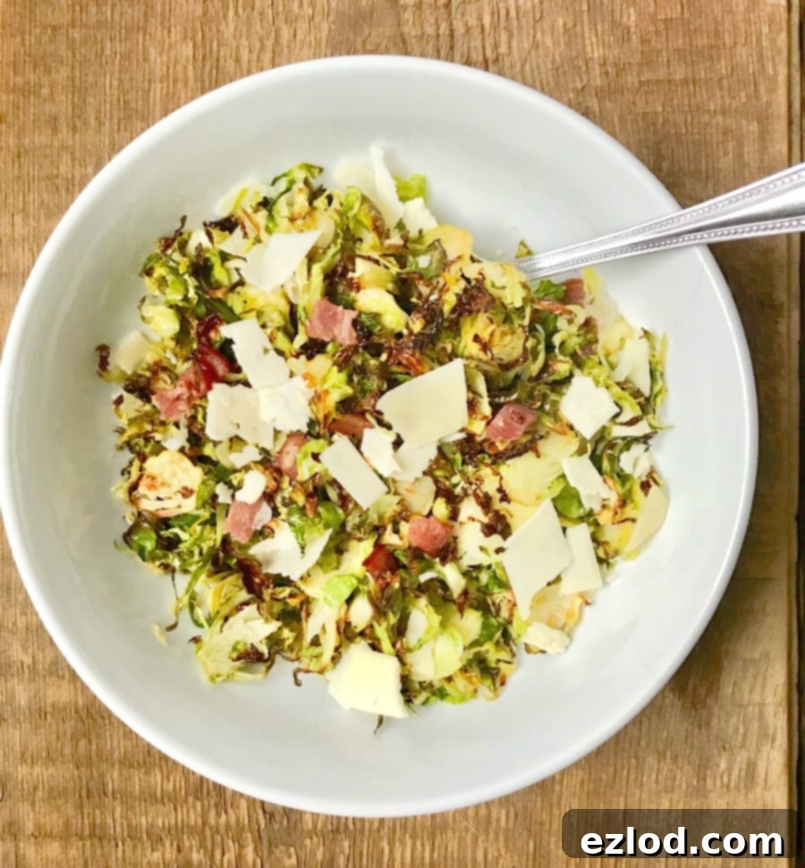 Overhead shot of a large white serving bowl with a silver spoon on a wooden surface. In the bowl is crispy shaved brussels sprouts topped with diced turkey bacon, shaved parmesan cheese, and a balsamic vinegar reduction.