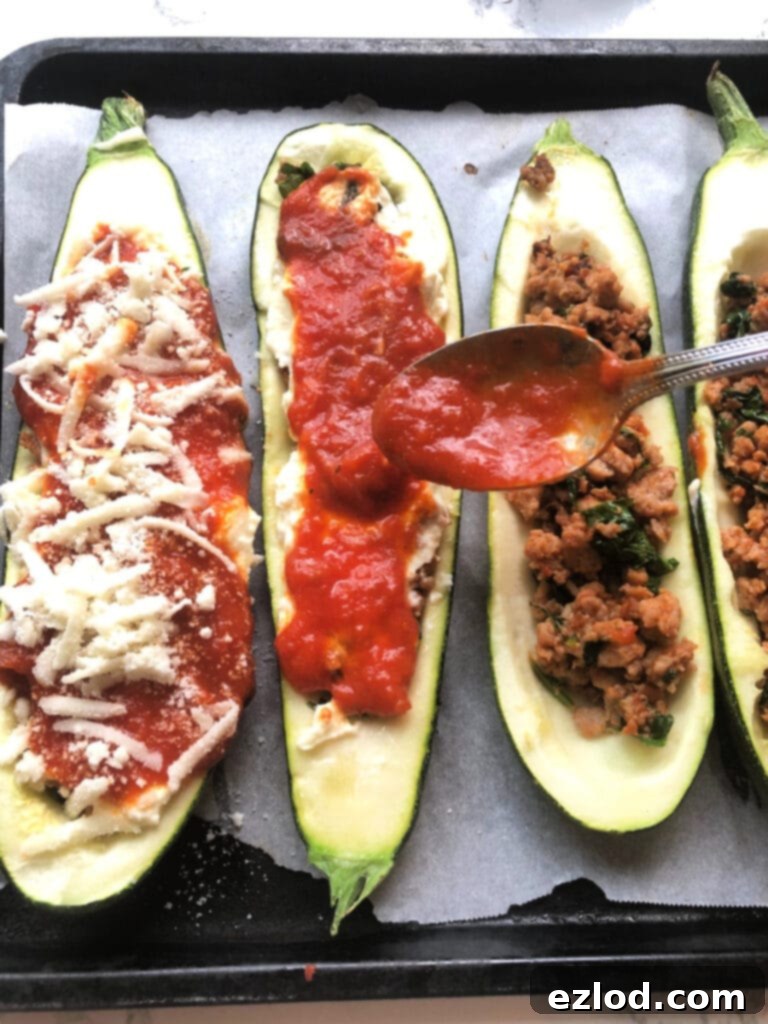 A hand spoons vibrant red marinara sauce over beautifully assembled Lasagna Stuffed Zucchini Boats, showcasing the final layer of flavor being added before baking to perfection. The layers of meat and cheese are visible beneath.