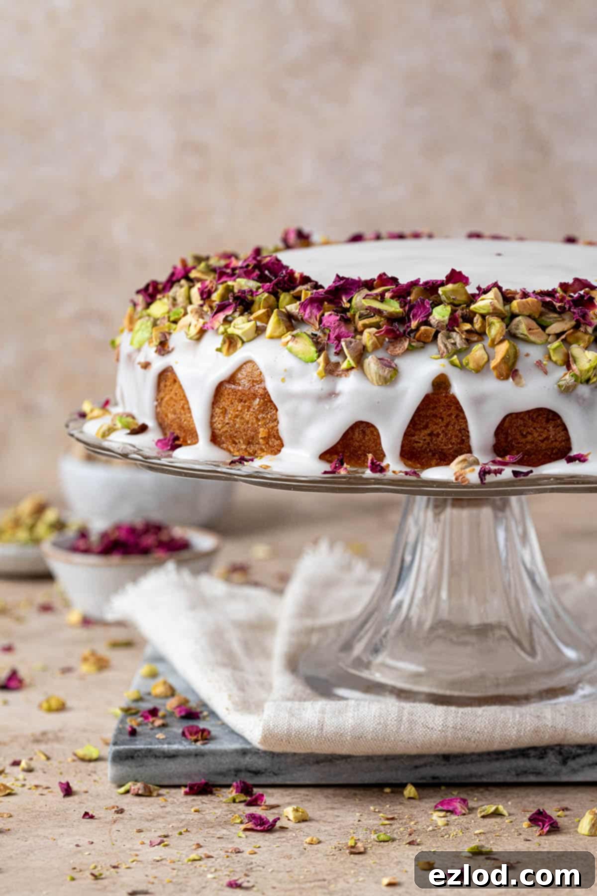 Vegan Persian love cake topped with pistachios and rose petals on a glass cake stand.