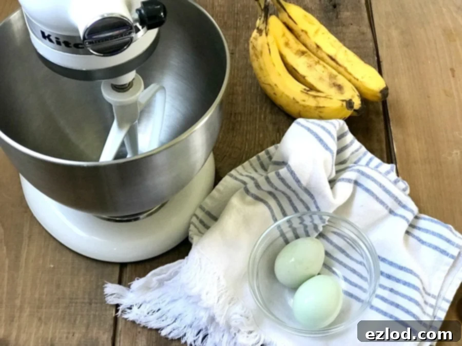 Overhead photo of two eggs in a glass bowl on a blue and white striped kitchen towel, three bananas, and a stand mixer on a wooden surface.