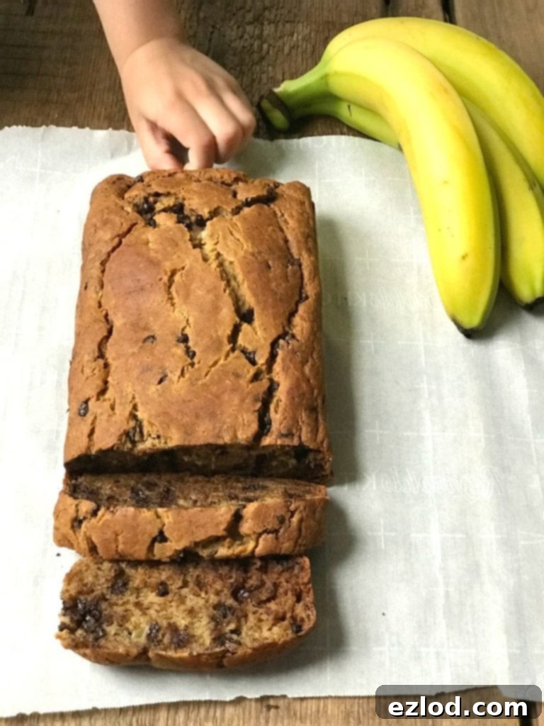 Overhead photo of a loaf of banana bread with a few slices already cut. There is a little hand sneaking in trying to take a chocolate chip from the bread. Three bananas are off to the side of the parchment lined wooden surface.