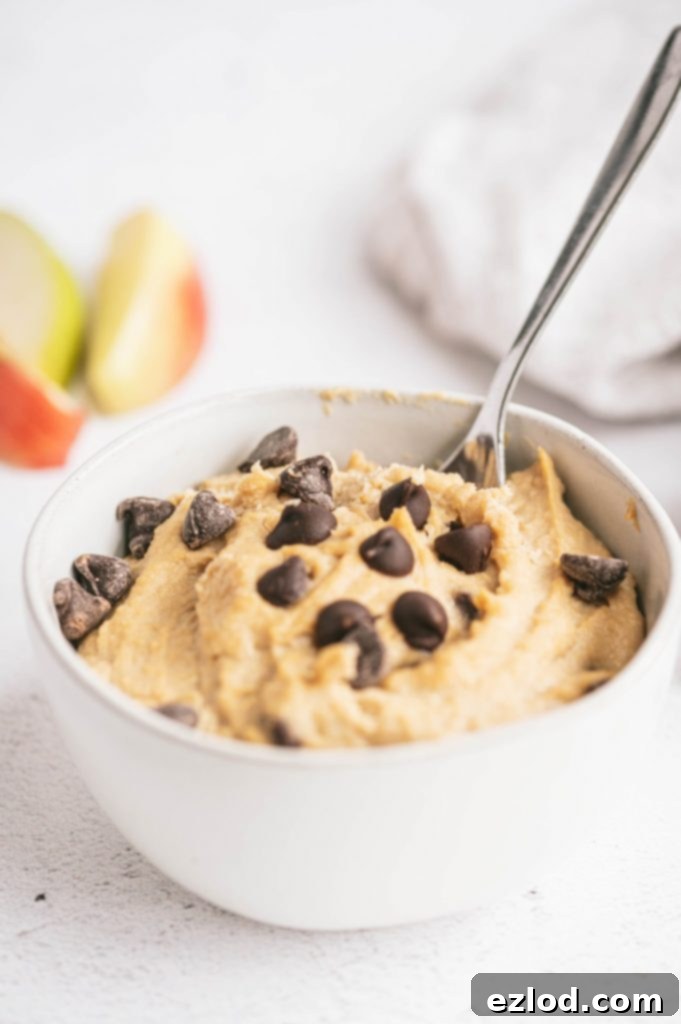 A bowl of healthy cookie dough with a spoon and several apples in the background, highlighting the creamy texture and chocolate chips.
