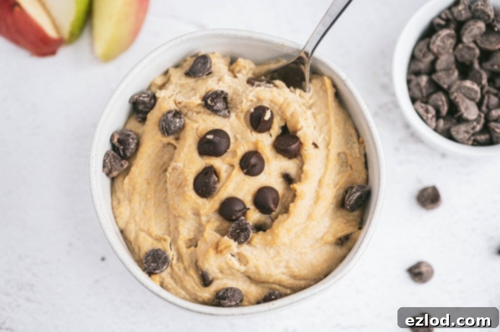 A small bowl of chickpea cookie dough dip, garnished with extra chocolate chips, alongside fresh apple slices for dipping.