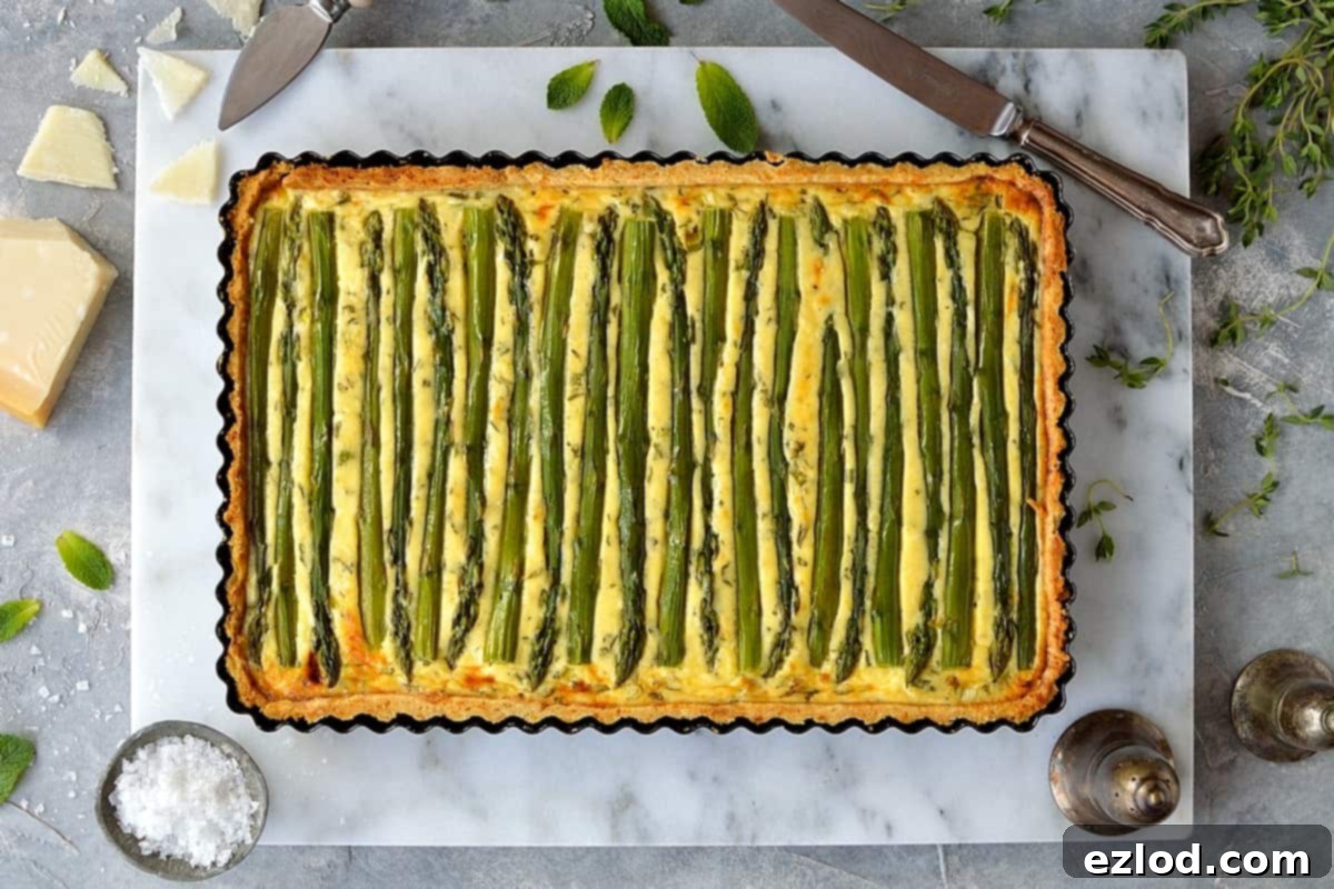 A freshly baked Asparagus, Pea, and Feta Cheese Tart (Quiche) on a white platter with a single slice removed, ready to be served. It's garnished with fresh herbs.