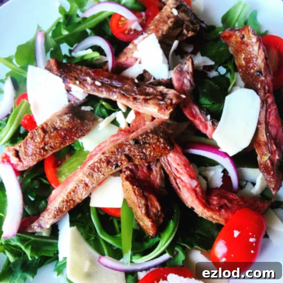 Fresh arugula salad topped with halved grape tomatoes, sliced red onions, generous shaved Parmesan cheese, and strips of perfectly grilled flank steak on a clean white plate. A healthy steak salad.