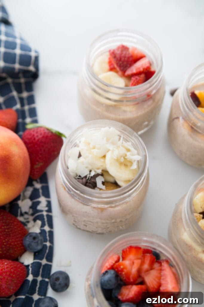 Five meal prep Overnight Oats with various toppings on a marble backdrop. A blue kitchen towel sits nearby and with berries and peaches on top