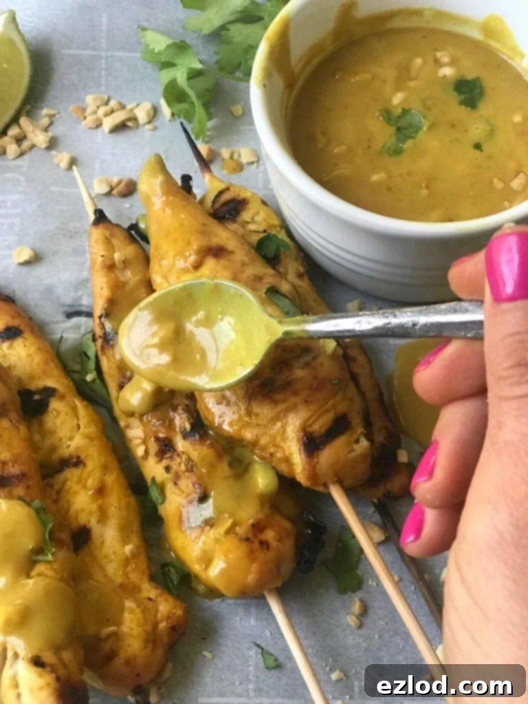 Chicken satay skewers drizzled with creamy peanut sauce dripping off spoon. A dish of the sauce is off to the side of the parchment lined tray.