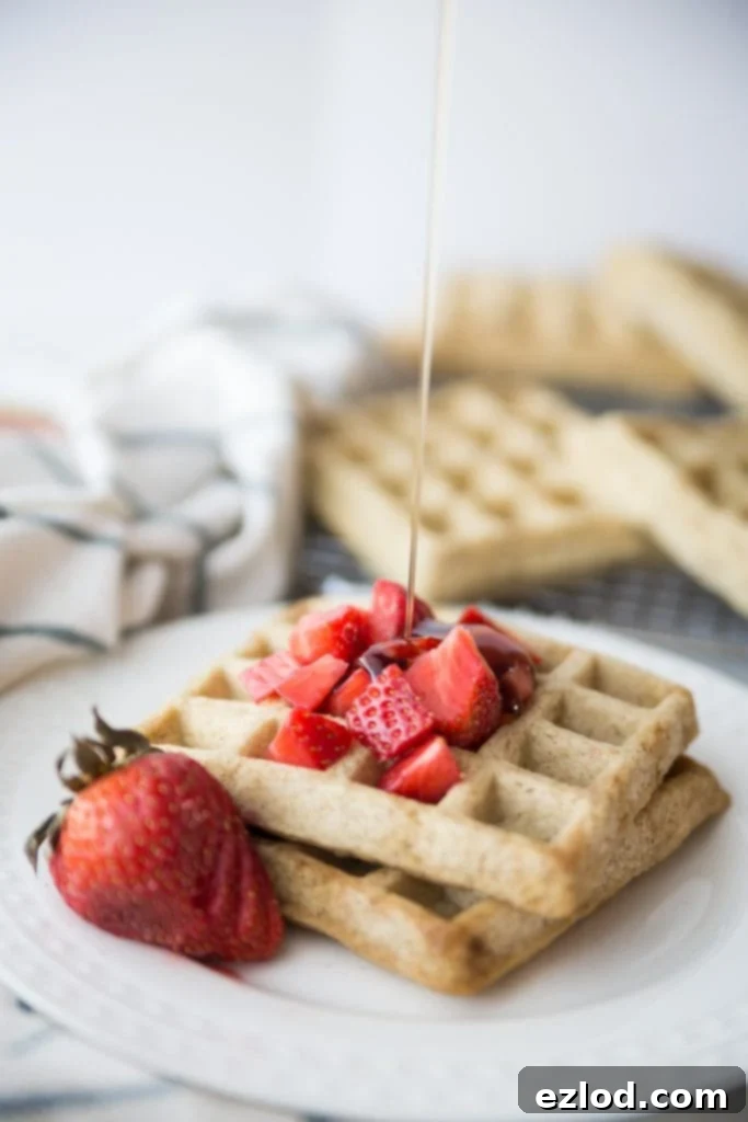 Two square homemade waffles on a white plate topped with diced strawberries and a sliced strawberry for garnish. Maple syrup is being drizzled from above.