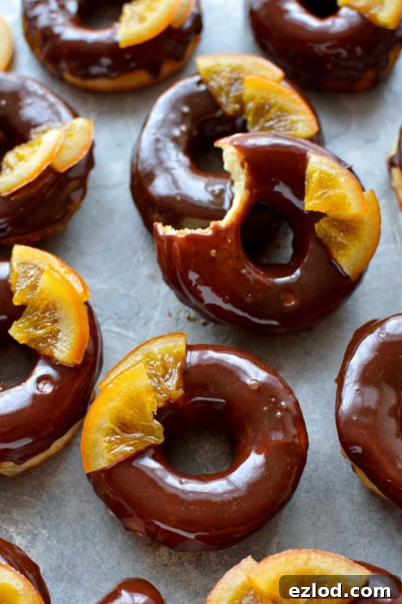 Stack of vegan chocolate orange doughnuts with glaze and candied orange on a white plate.