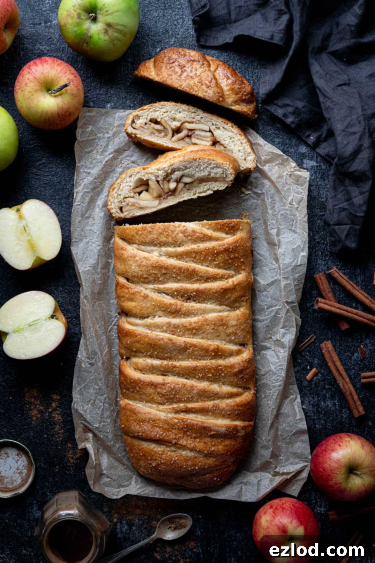 Spiced Apple Vegan Brioche 2 Vegan apple cinnamon brioche loaf on a black surface, surrounded by fresh apples and cinnamon sticks, highlighting its braided design and golden crust.