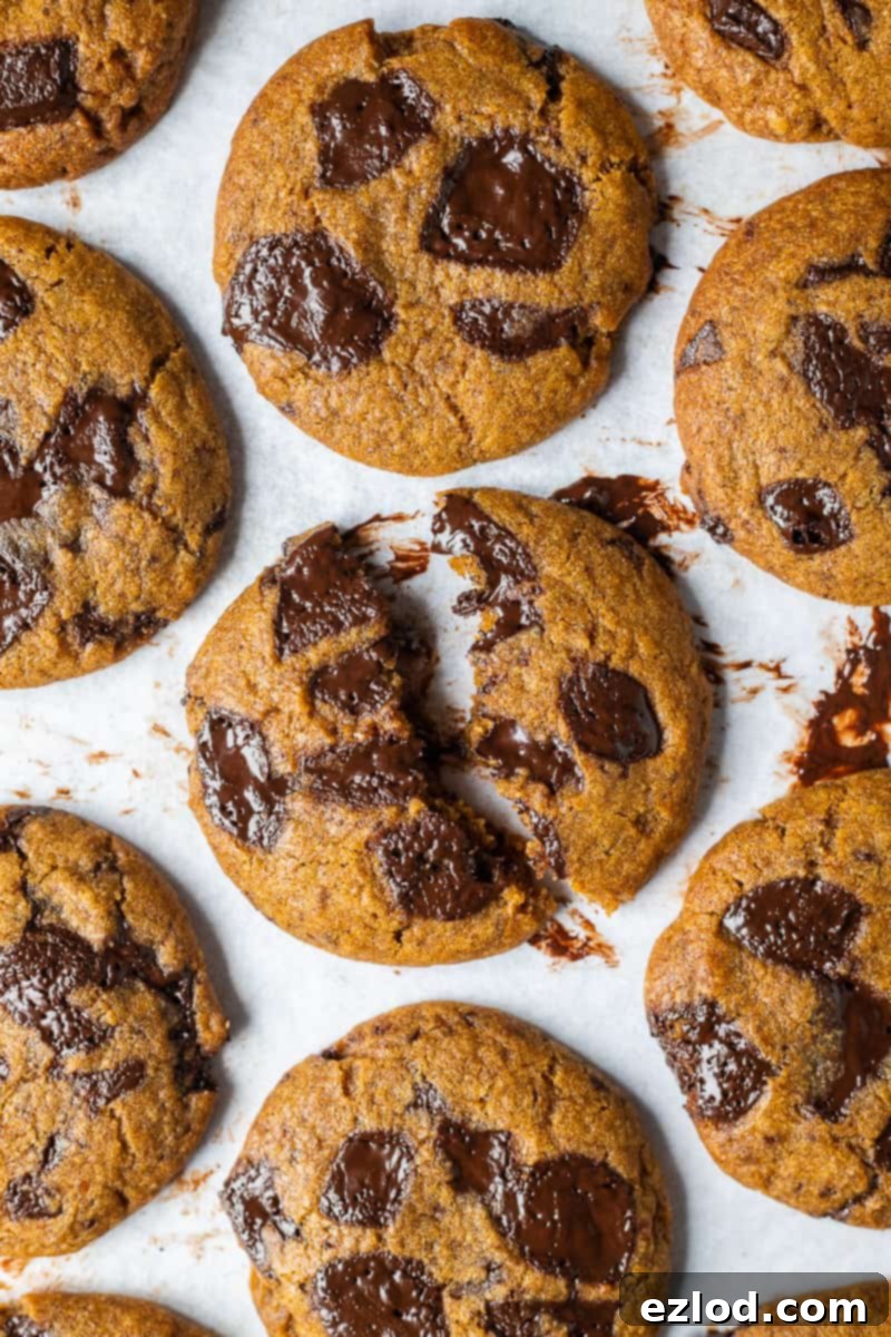 A close-up view of a vegan pumpkin spice cookie, broken in half to reveal its soft, moist, pumpkin-infused interior and melting chocolate chips. The texture looks perfectly chewy and inviting.