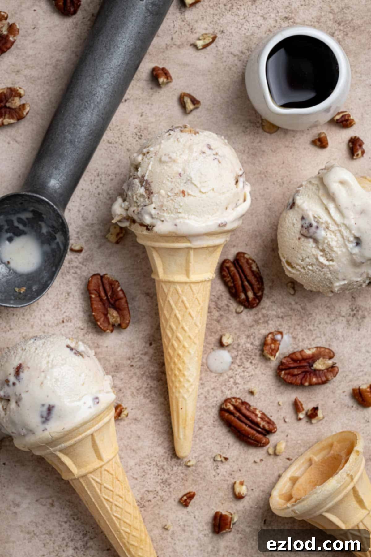 Vegan maple pecan ice cream cones with a jug of maple syrup, an ice cream scoop and pecans on a brown surface.