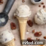 Vegan maple pecan ice cream cones with a jug of maple syrup, an ice cream scoop and pecans on a brown surface.