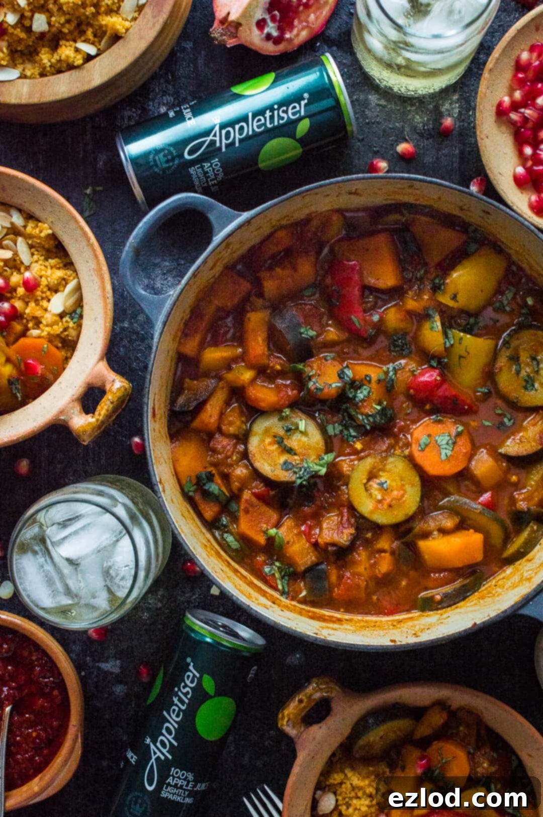 A vibrant flatlay of the wholesome vegan vegetable tagine, adorned with fresh herbs, served alongside a bowl of almond and chickpea couscous. A sparkling Appletiser beverage completes this perfect healthy meal arrangement.