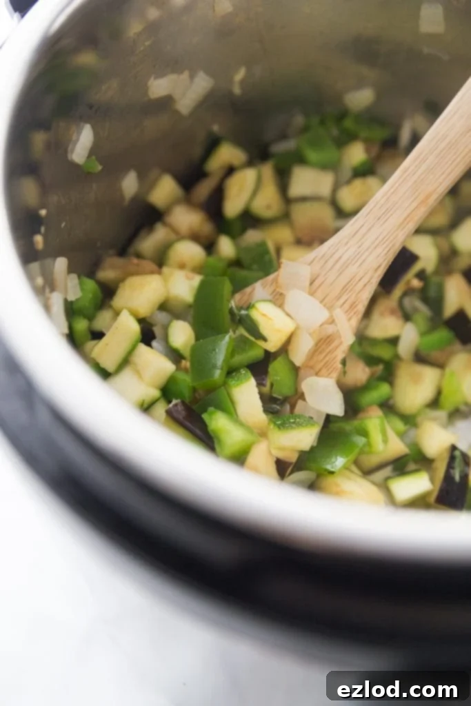 A close-up of chopped zucchini, bell peppers, and onions being sautéed in the stainless steel pot of an Instant Pot, with a wooden spoon resting on the side.