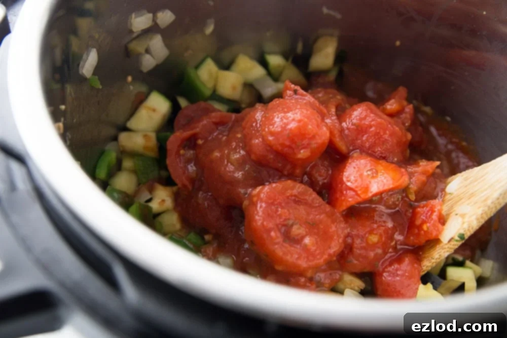 A top-down view of an Instant Pot filled with sautéed vegetables, with a pile of canned stewed tomatoes freshly added on top, ready to be stirred. A wooden spoon rests nearby.