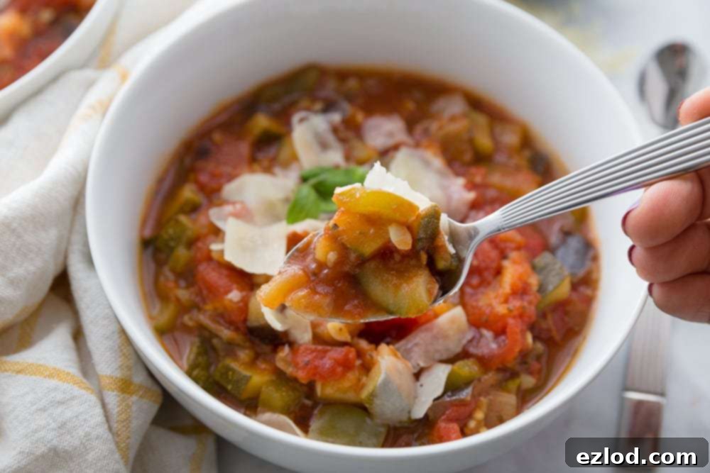 A wooden spoon holds a perfect bite of Instant Pot Ratatouille, with a white bowl of ratatouille topped with Parmesan and fresh basil blurred in the background, ready for eating.