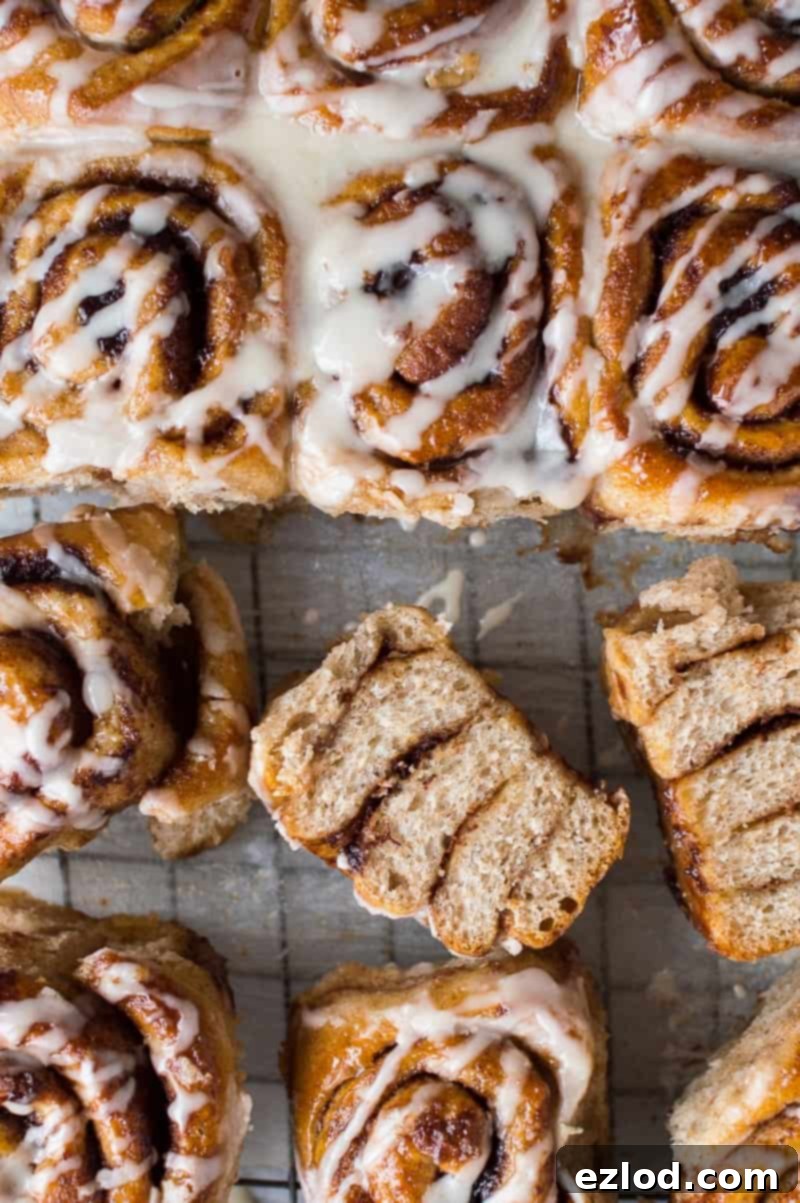 A stunning top-down view of freshly baked vegan wholemeal maple cinnamon rolls, generously coated with a luscious maple glaze, with one bun thoughtfully sliced open to reveal its irresistible sticky, swirled filling.