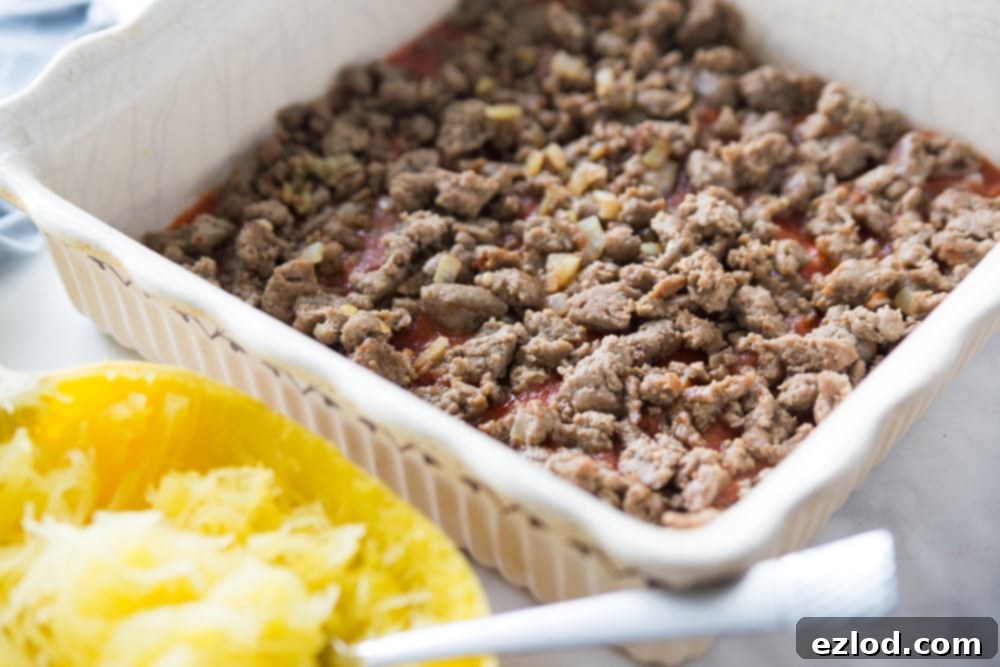 Half of a cooked spaghetti squash sitting next to a square white baking dish filled with cooked ground meat and tomatoes, showcasing a healthy meal.
