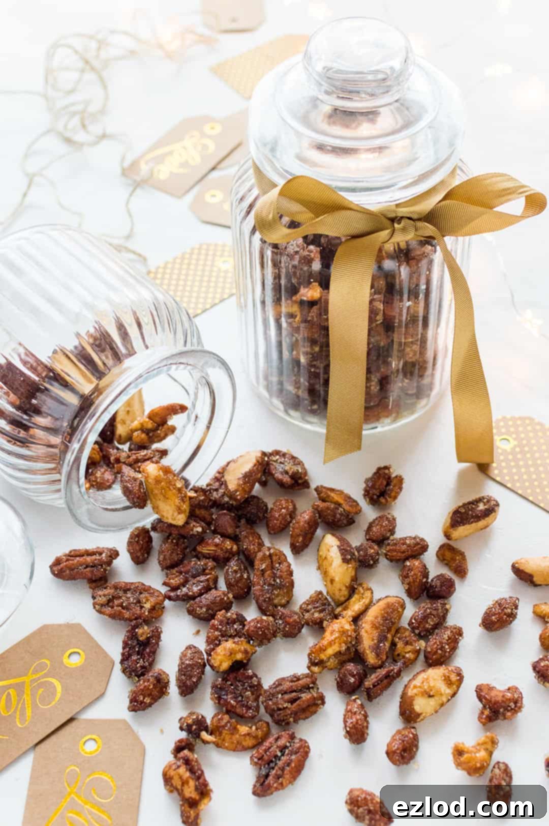 Spiced honey roasted mixed nuts tumbling out of a glass cookie jar with festive ribbon and gift tags, ready for gifting.