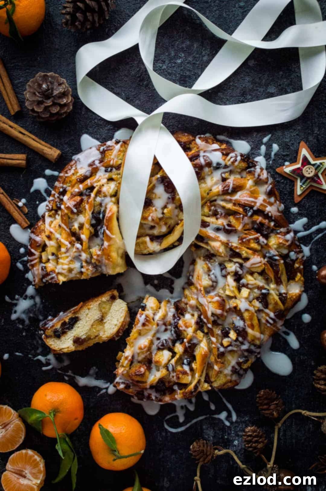mincemeat, marzipan and apple bread wreath on a black background with a slice taken out surrounded by clementines, spices, ribbon and pine cones.