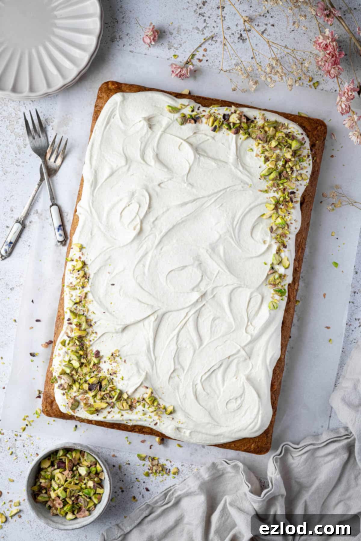 The assembled carrot, pineapple and pistachio cake with dried flowers, plates, forks and a bowl of pistachios for decoration.