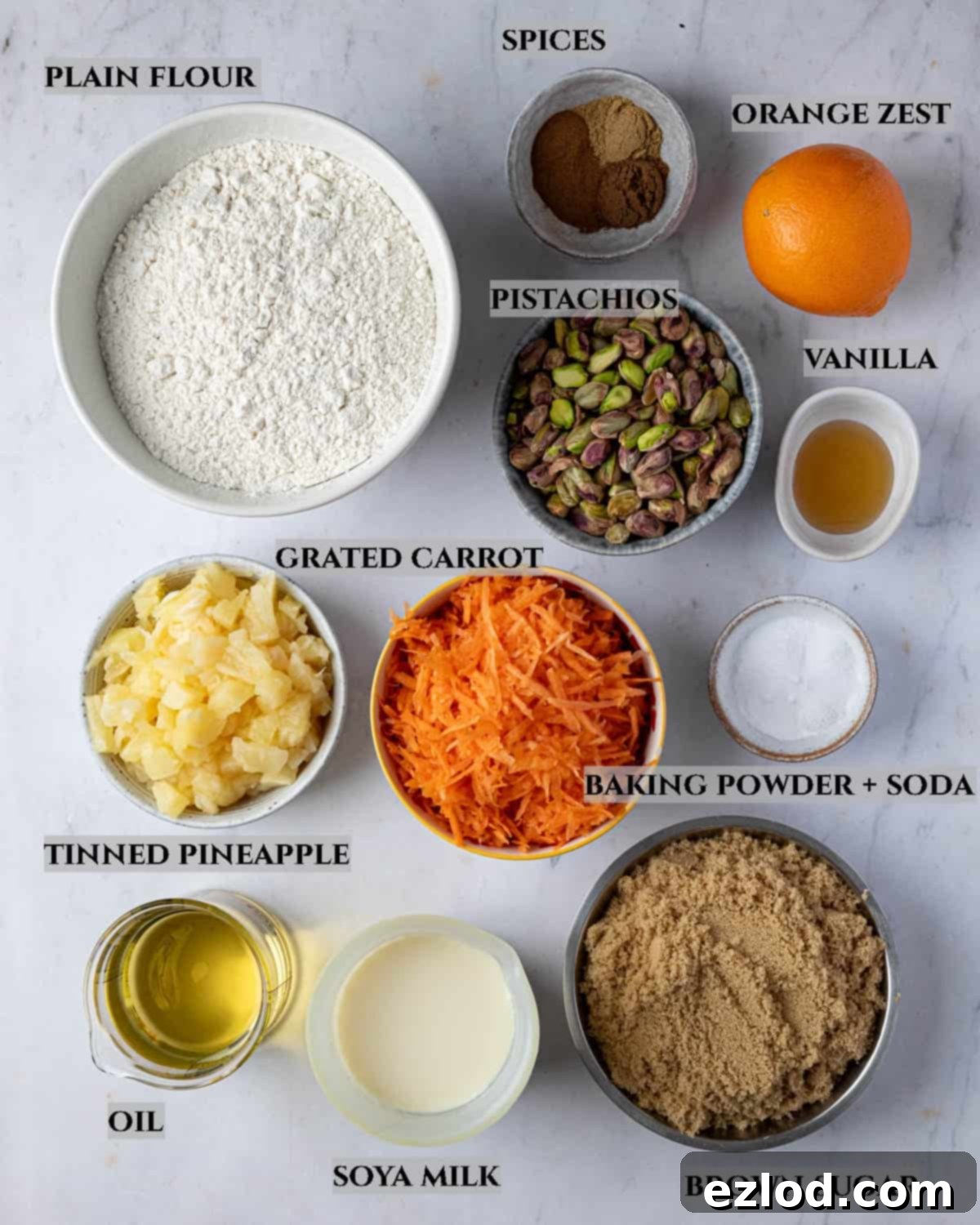 A labelled photo of the ingredients needed to make vegan pineapple and pistachio carrot cake, showcasing fresh carrots, a can of pineapple, and pistachios.