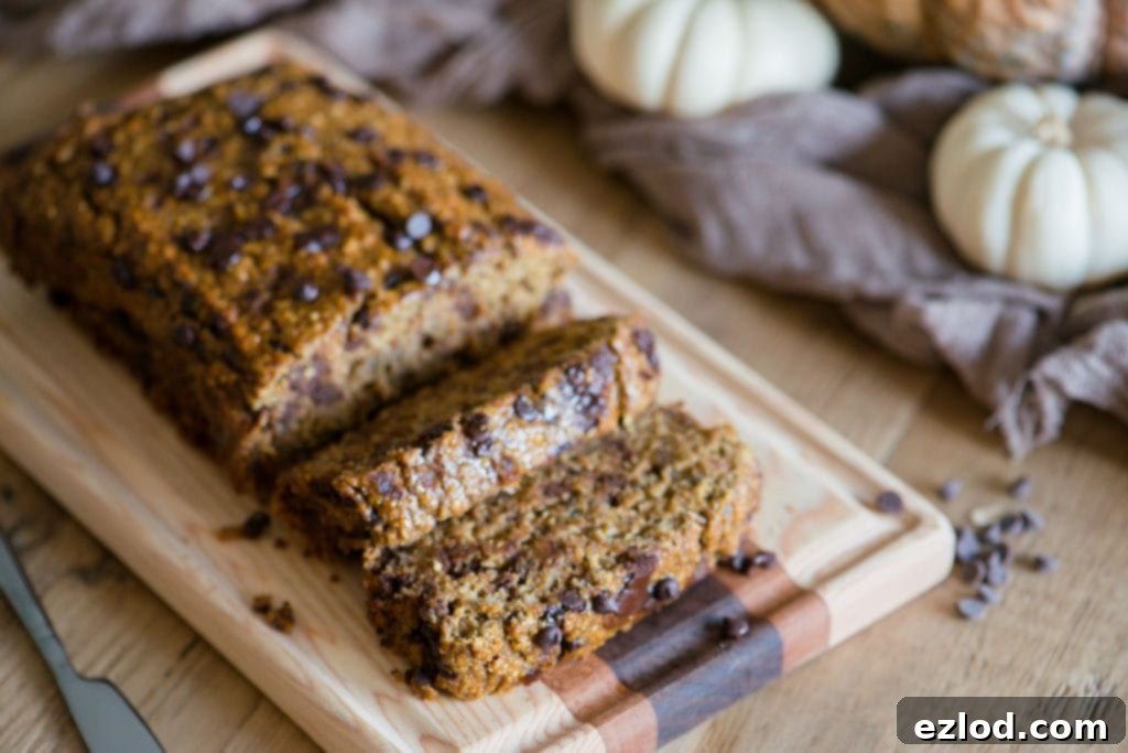 Sliced Pumpkin Oatmeal banana bread with chocolate chips laying on a wooden cutting board. The light colored wood table is adorned with white and orange pumpkins and a light brown piece of fabric. A small knife sits beside the board. 