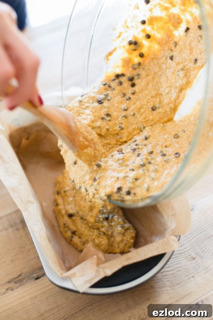 A woman's hand is shown using a wooden spoon to pour oatmeal banana pumpkin bread into a loaf pan lined with parchment paper. 