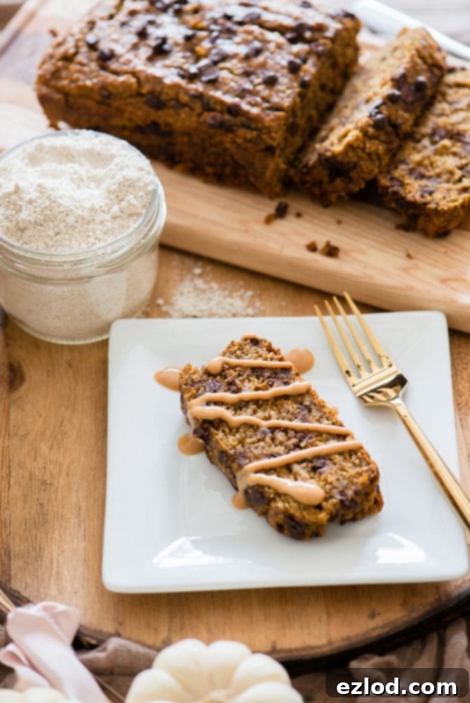 A loaf of Pumpkin Banana Oatmeal Bread sits on a wooden tray. Next to the bread is a small mason jar of oat flour and a plate with a slice of the bread with peanut butter drizzled on it. 