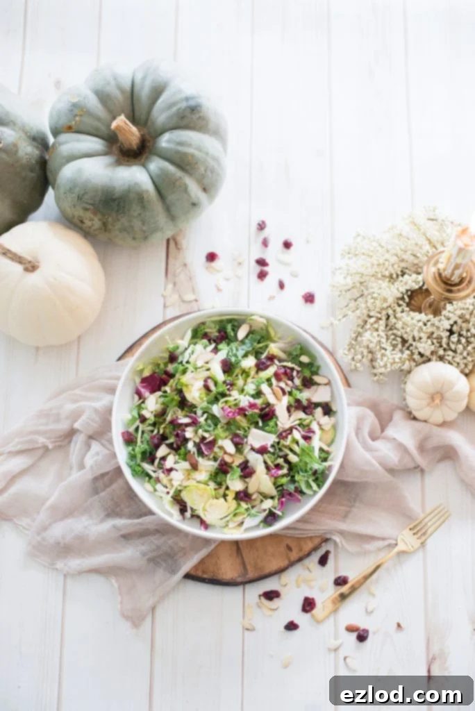 Overhead photo of Maple Cider Holiday Salad with festive fall decor. White, cream, and muted blue decorations surround the white serving bowl full of greens, dried cranberries, and sliced almonds.