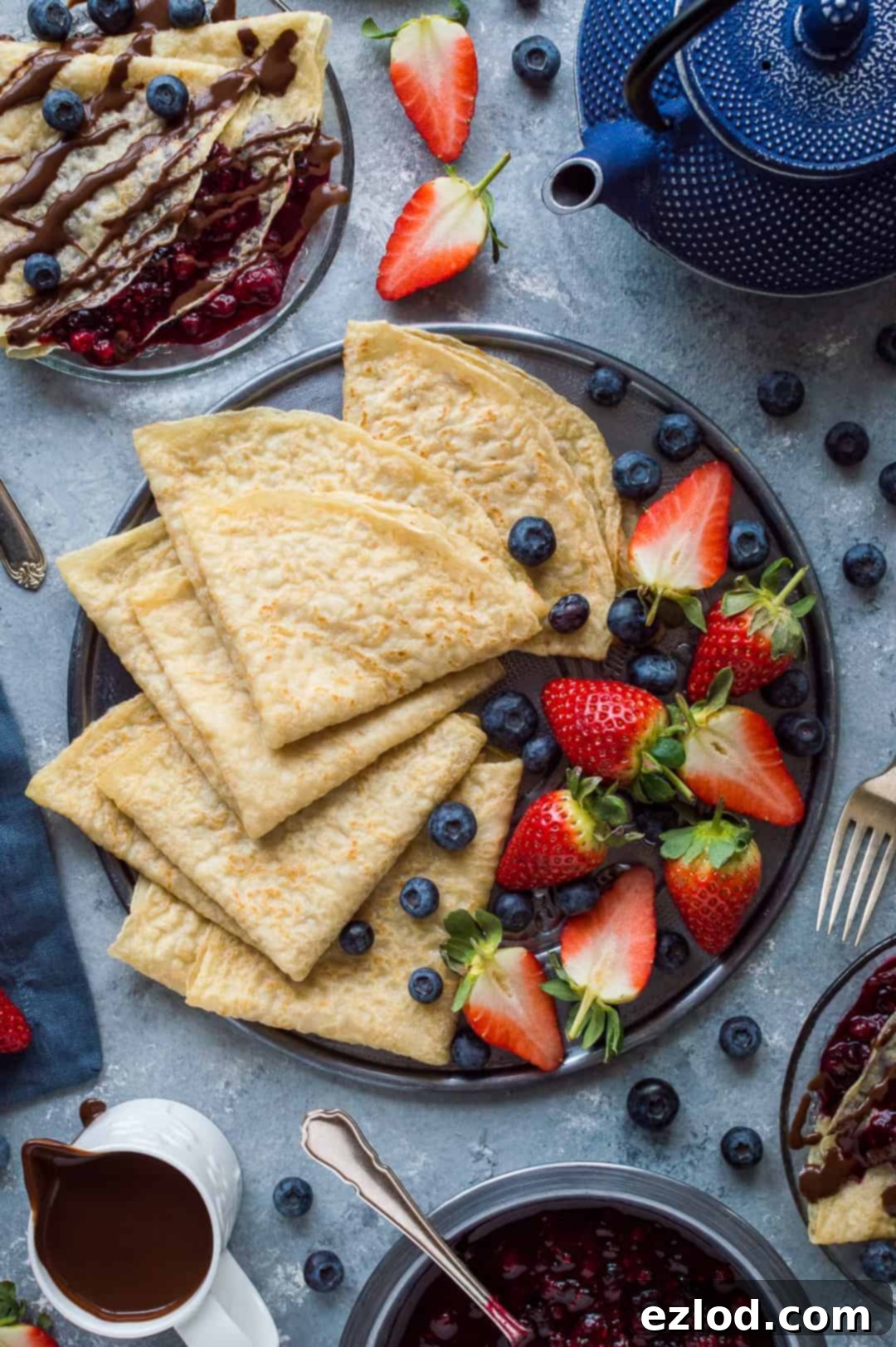 Vegan crepes folded into triangles on a metal plate with strawberries and blueberries, with a blue teapot, jug of chocolate sauce and plates of filled crepes.