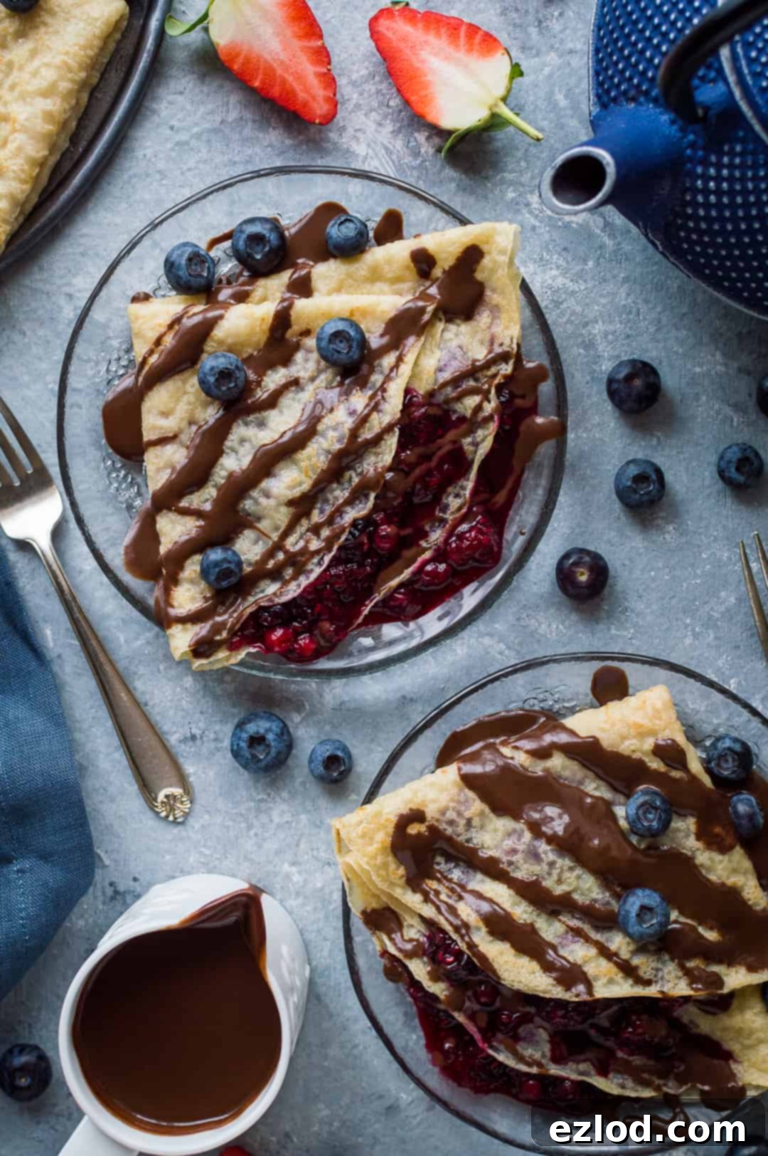 Two glass plates of pancakes filled with berry compote and drizzled with chocolate sauce and blueberries.