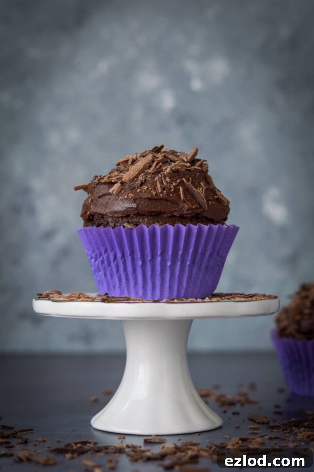 A single vegan chocolate cupcake, beautifully frosted and adorned with chocolate shavings, resting on a miniature cake stand.