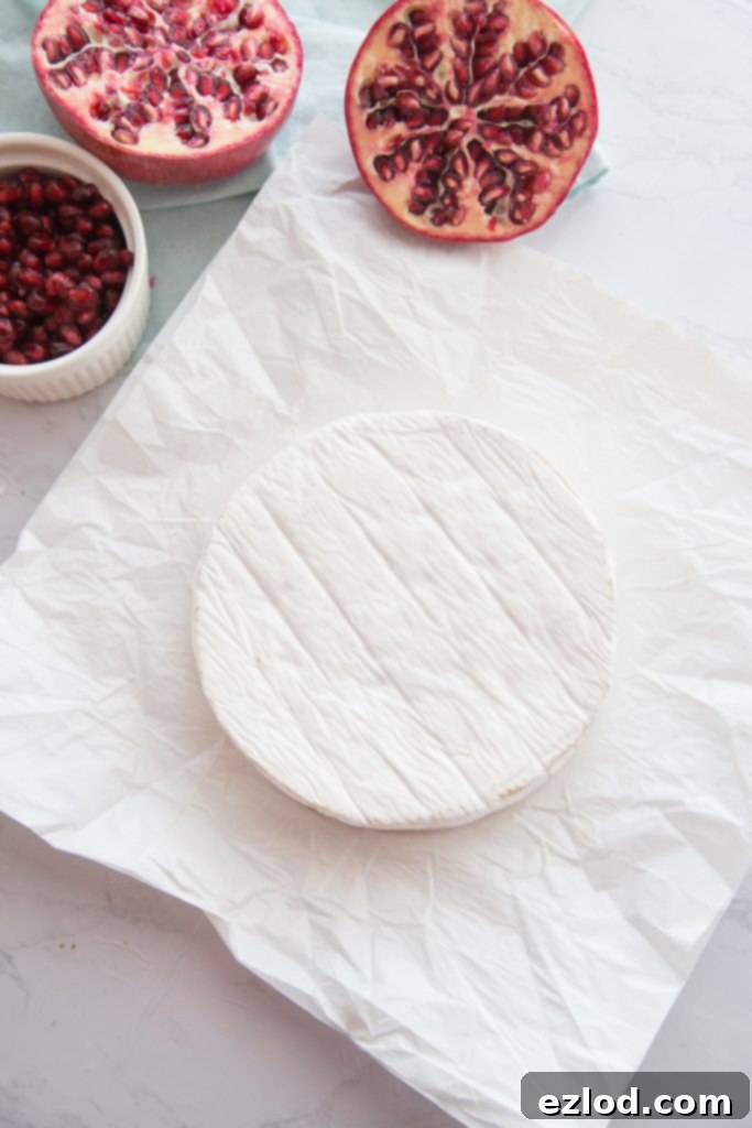 A brie wheel is unwrapped from it's white paper with a halved pomegranate on the side and a white bowl of pomegranate arils..