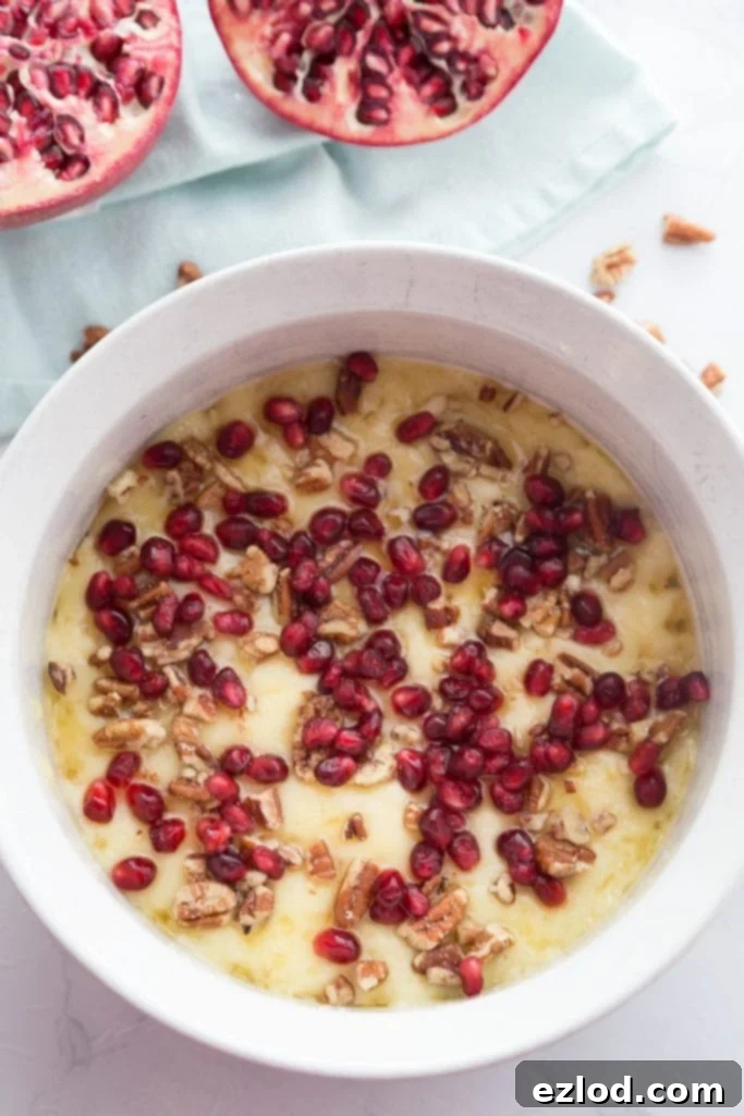 Overhead photo of a white ceramic bowl of melted brie with pomegranate seeds, honey and pecans. Two pomegranate halves sit nearby
