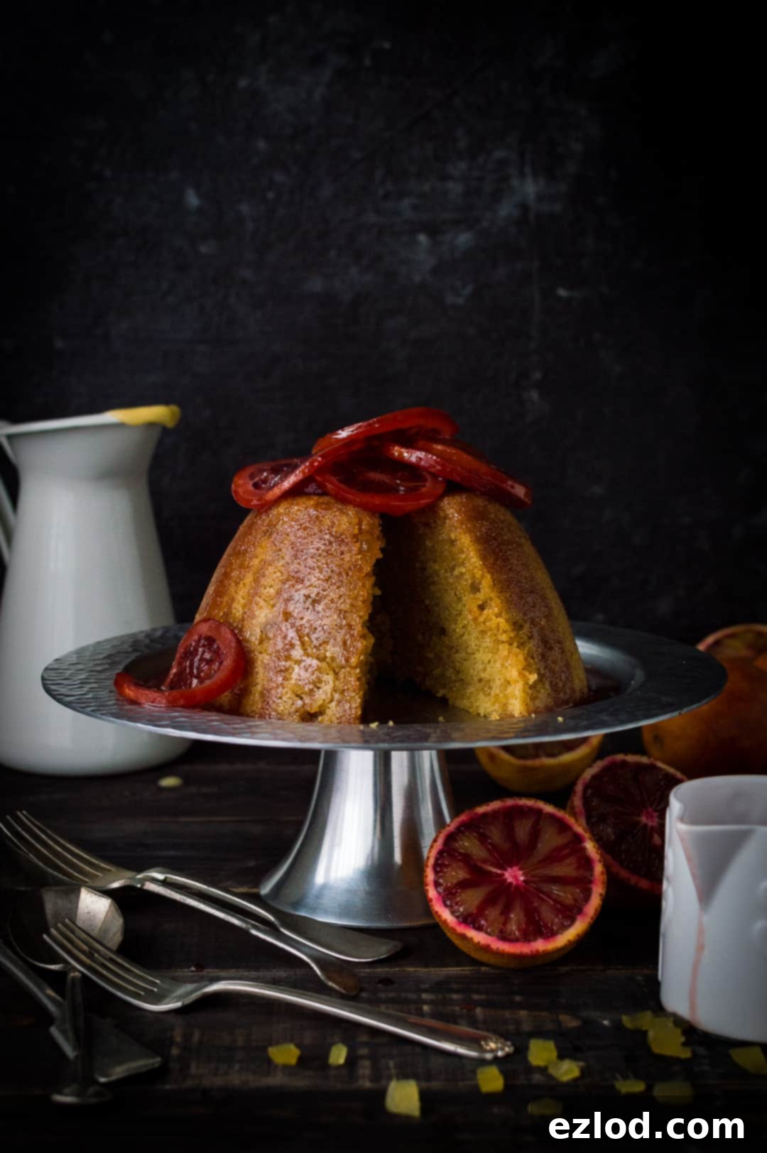 Elegant slice of blood orange and ginger steamed pudding, presented on a silver cake stand with delicate candied orange garnish.