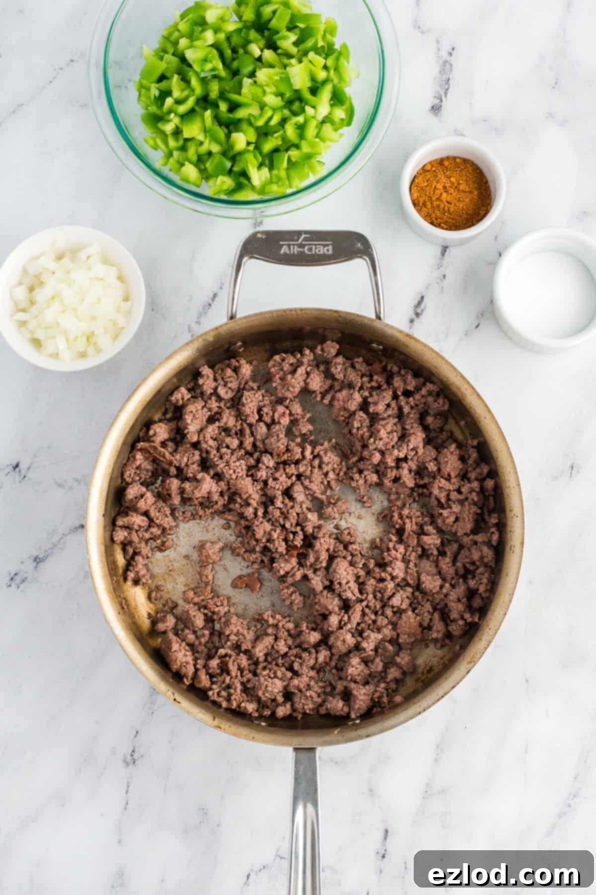 Overhead view of ground meat browning in a skillet, with bowls of chopped green bell peppers, onions, and seasonings arranged around it.