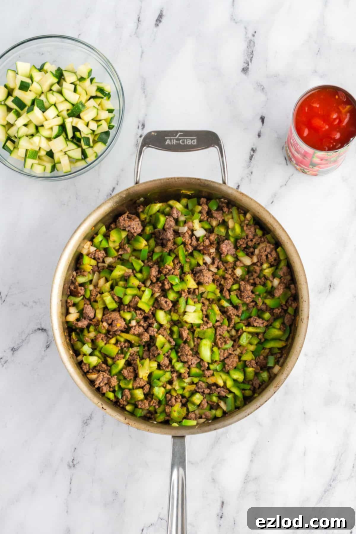 Overhead view of ground meat browning in a skillet, with a bowl of zucchini and a can of diced tomatoes nearby.