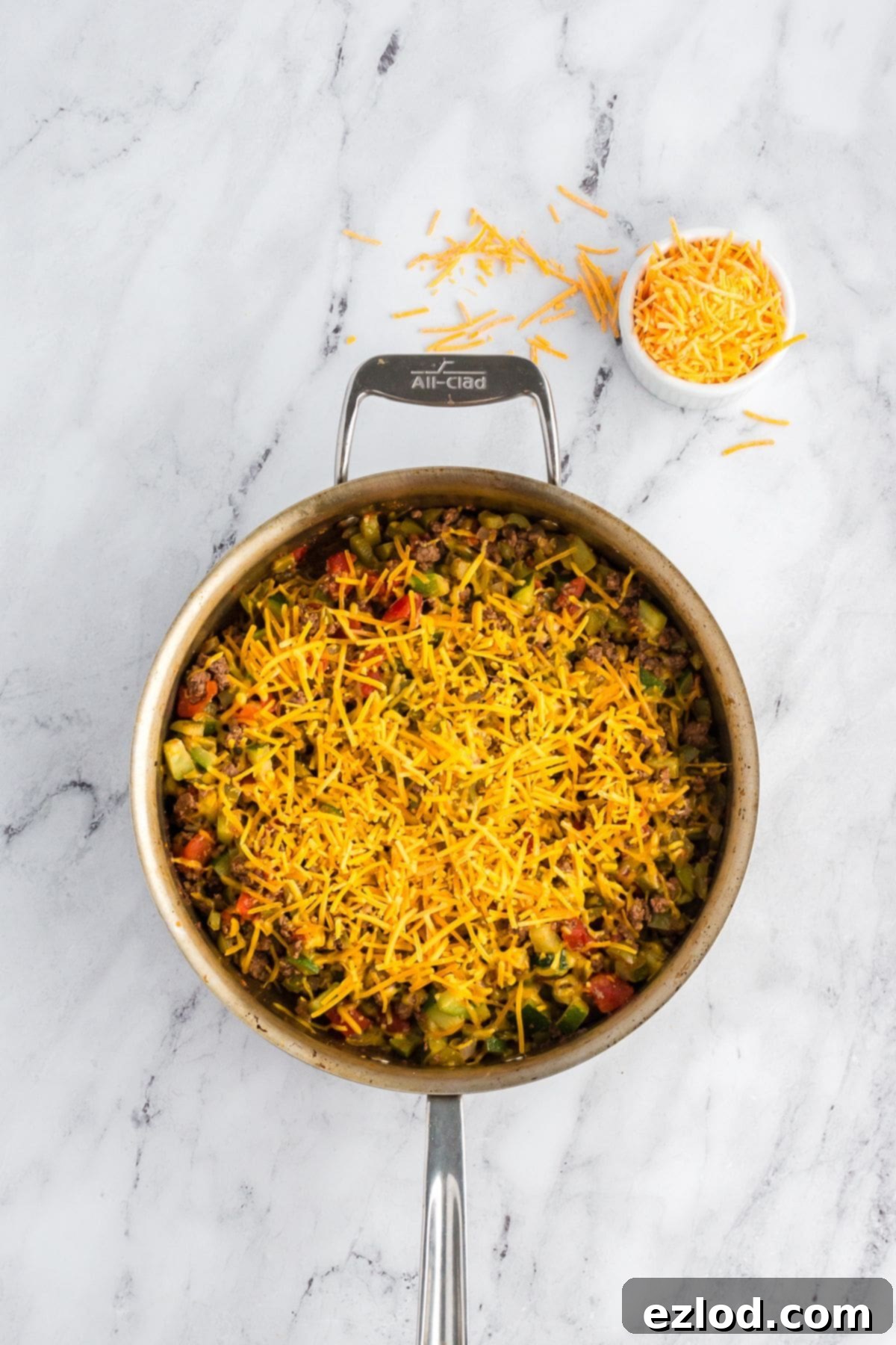 Overhead view of a skillet with taco zucchini mixture topped with shredded cheese before melting, with cheese scattered on the marble surface.