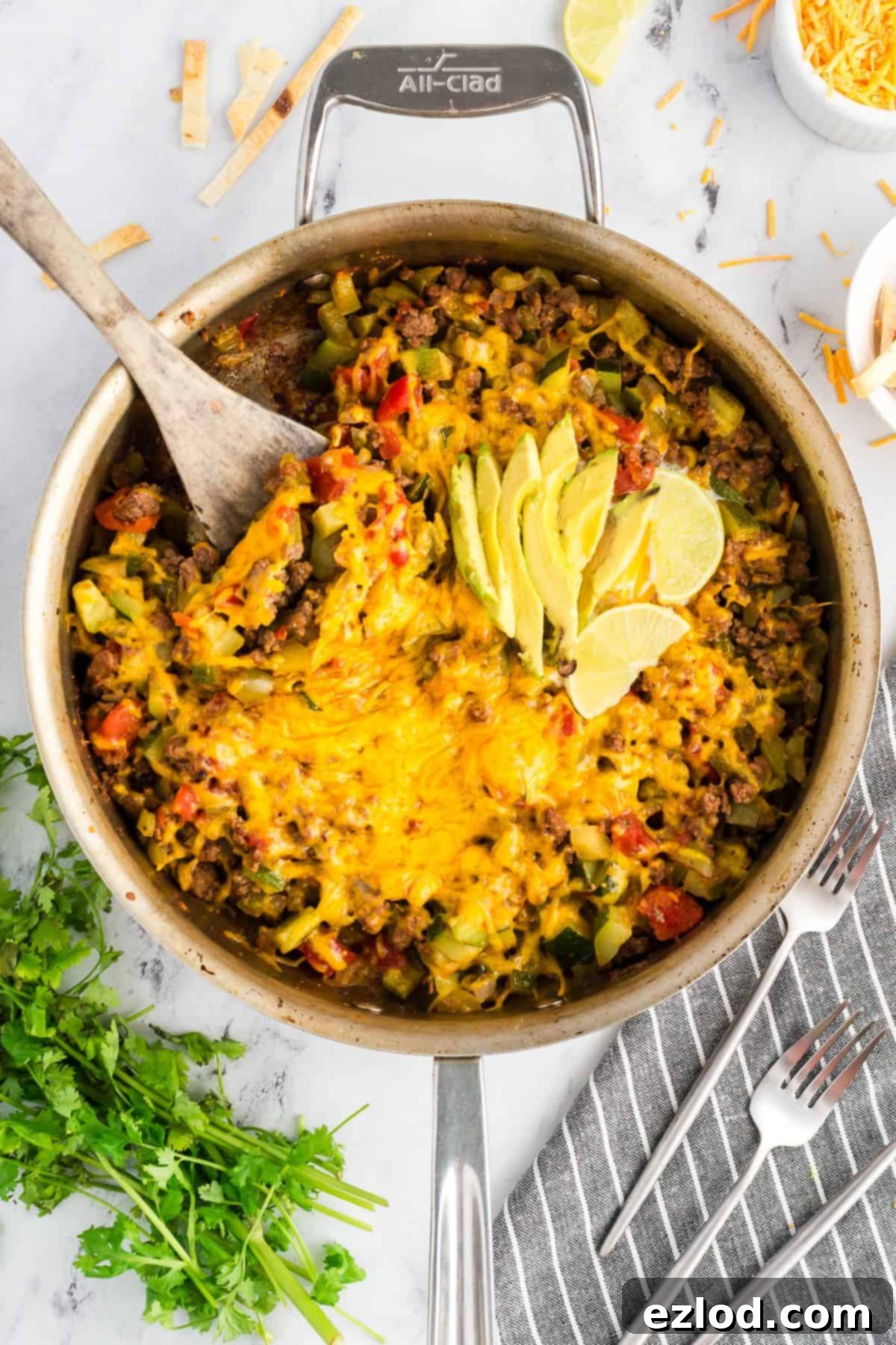 Overhead view of a colorful taco zucchini skillet in a pan, topped with melted cheese and sliced avocado, with lime wedges.