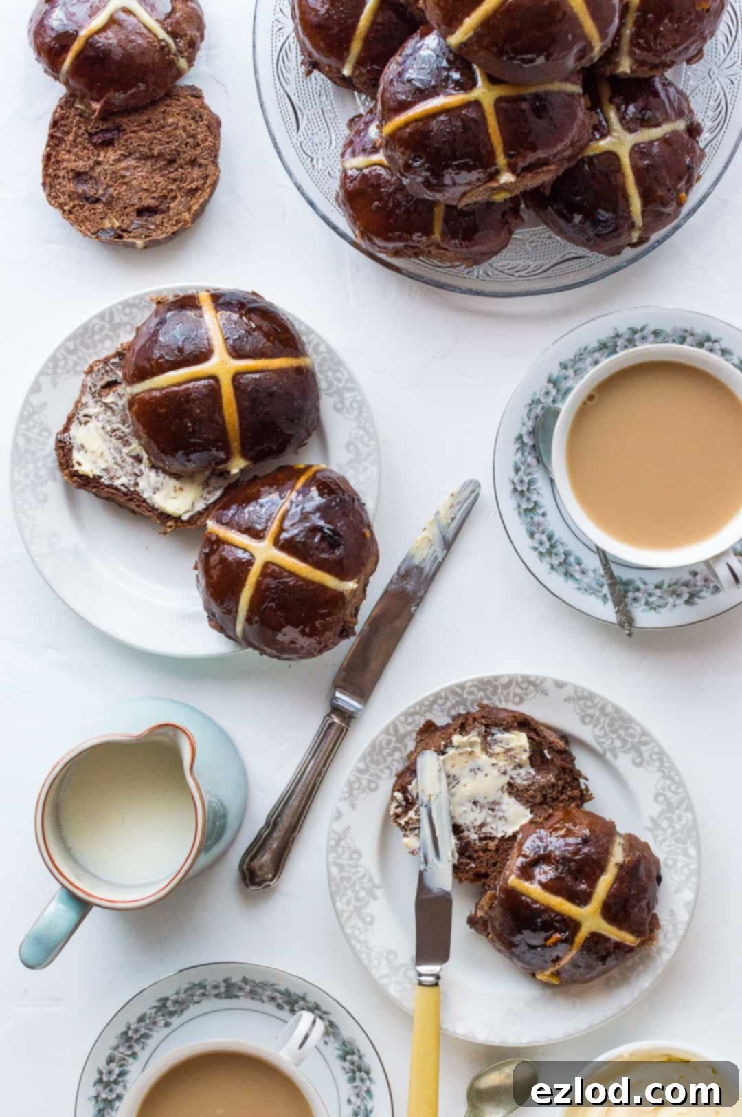 Freshly baked chocolate orange hot cross buns neatly arranged on a pristine white surface, accompanied by a small dish of butter and two steaming cups of tea, ready for serving.