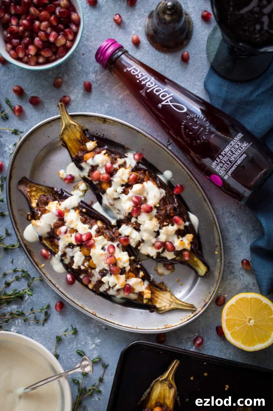 Stuffed eggplant with chickpeas, lentils, feta and lemon tahini sauce with a bottle of Appletiser on a grey background.