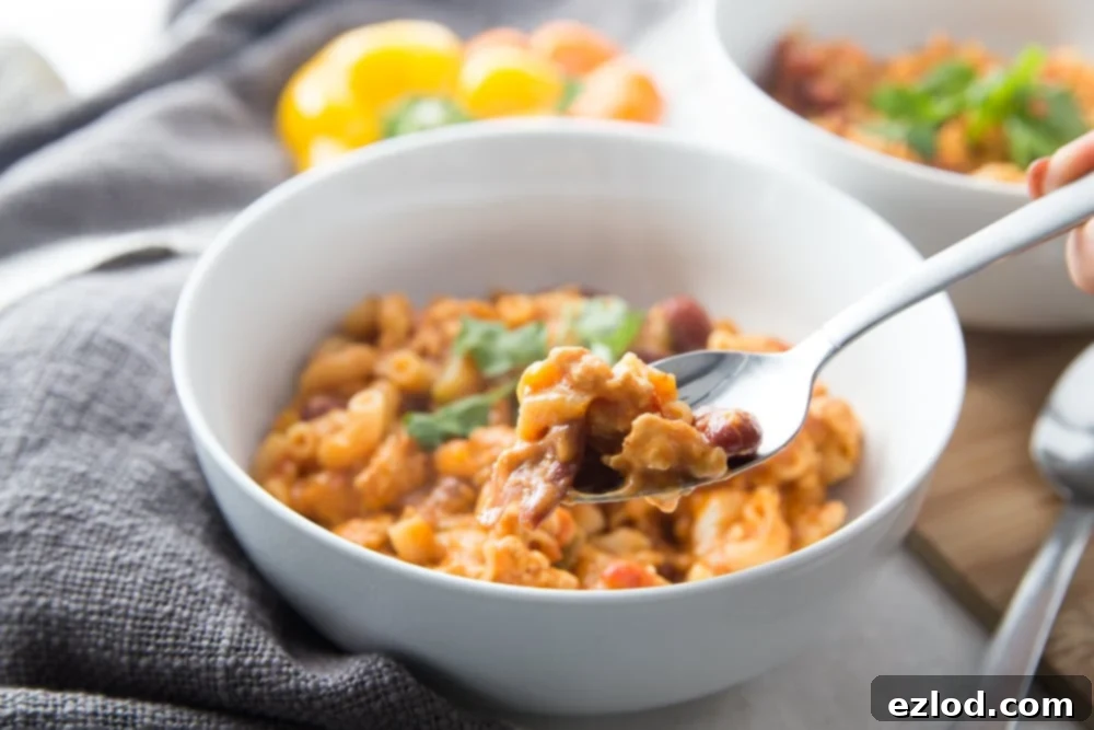 White bowl with healthy instant pot chili mac topped with cilantro. A hand is holding a spoon and about to take a bite. There is a second bowl seen in the background and some bell peppers sitting nearby.