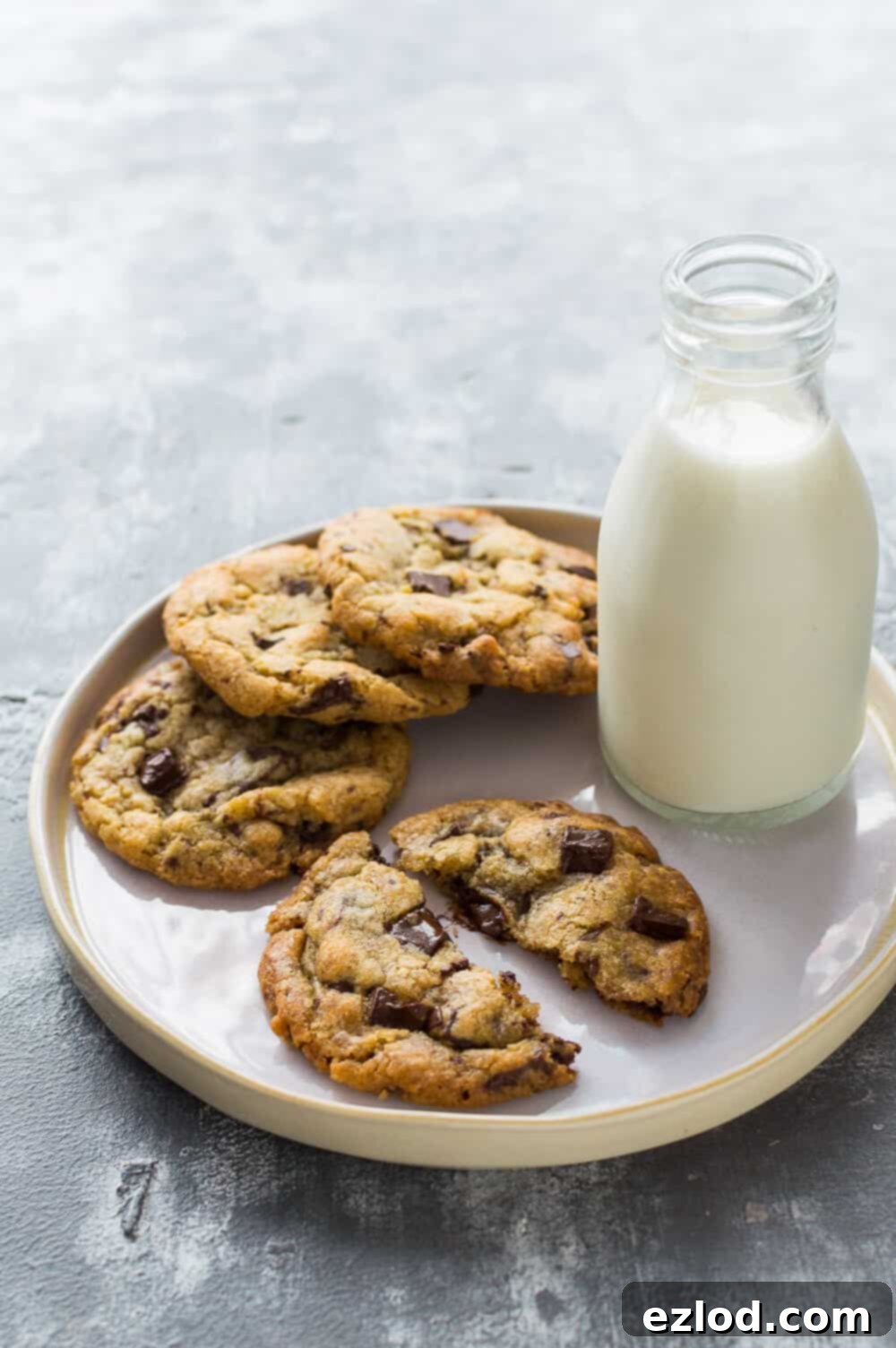 Photo of a white plate of vegan chocolate chip cookies with a glass bottle of almond milk on a grey background