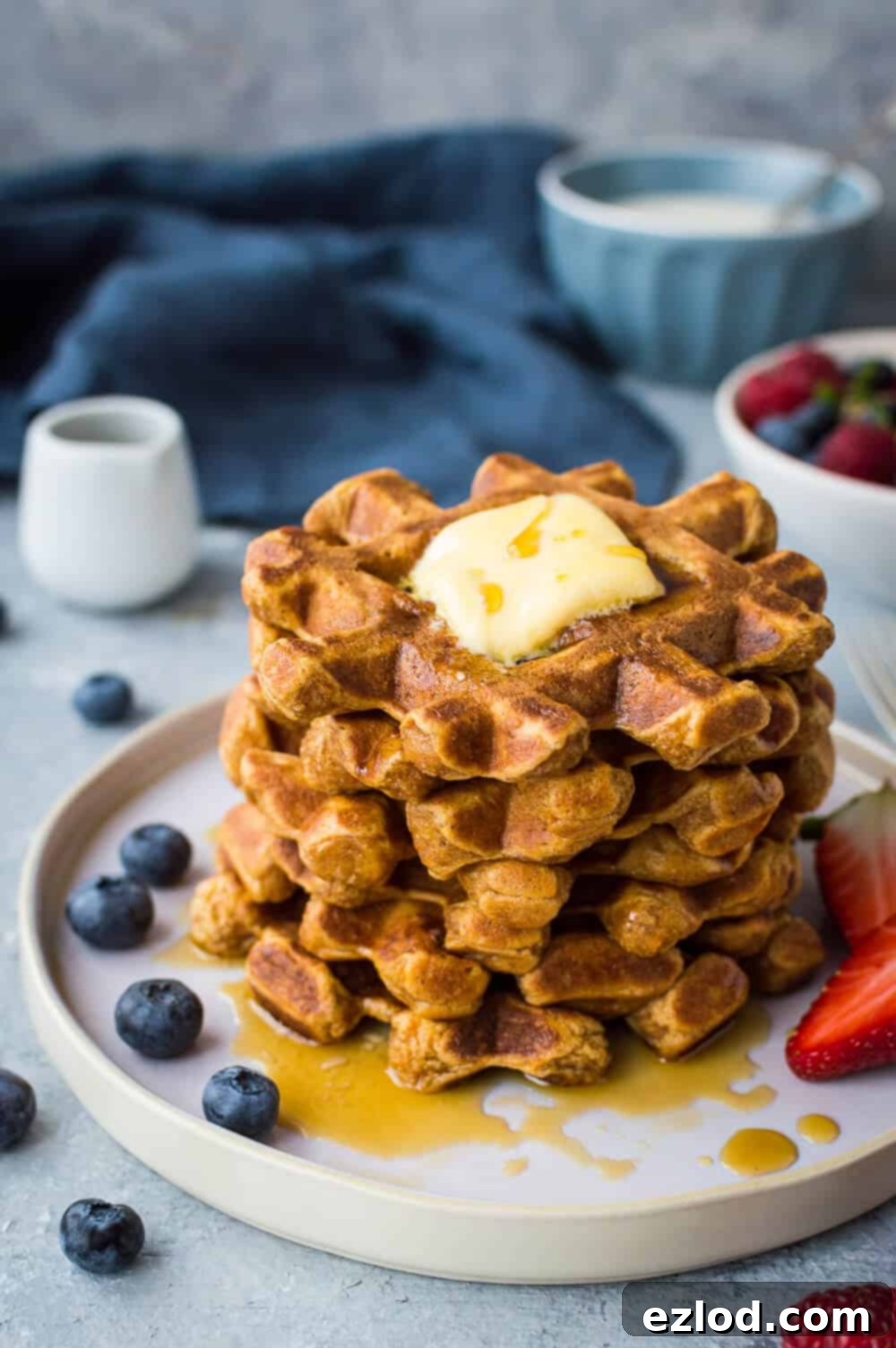 A visually appealing stack of golden-orange sweet potato waffles, elegantly served with a pat of melting butter, a generous pour of maple syrup, and a vibrant scattering of fresh berries.