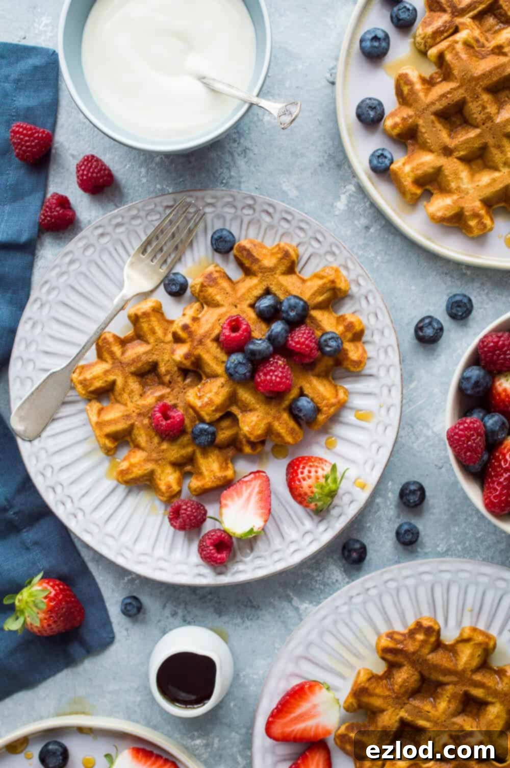 Three individual plates showcasing beautifully spiced sweet potato waffles, each garnished with maple syrup and fresh berries, artfully arranged on a stylish grey background.